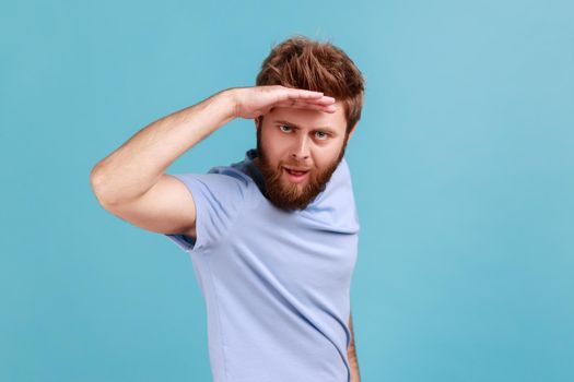 Portrait of handsome young adult bearded man looking far away at distance with hand over head, sees something pleasant with flirting expression. Indoor studio shot isolated on blue background.