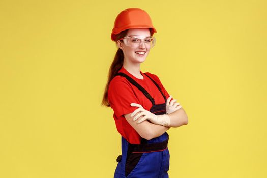 Side view of positive worker woman standing with crossed arms and looking at camera with confident expression, wearing overalls and helmet. Indoor studio shot isolated on yellow background.