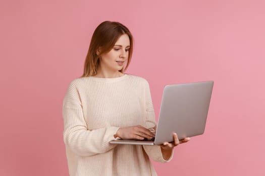 Portrait of positive beautiful young adult blond woman working on laptop compute, looking at display with smile, wearing white sweater. Indoor studio shot isolated on pink background.