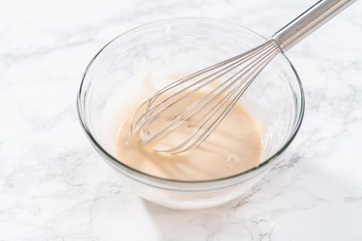 Lemon glaze. Mixing ingredients with a hand whisk in a glass mixing bowl to make lemon glaze.