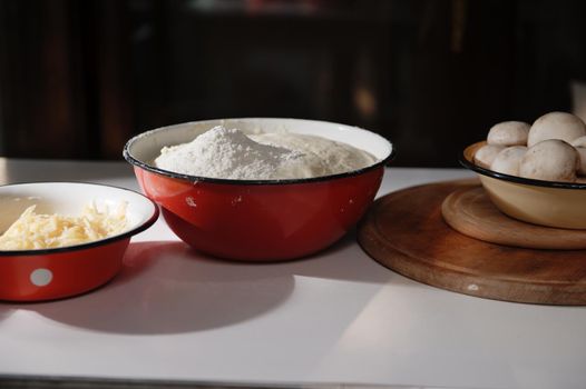 Enamel bowls with yeast rising dough, sprinkled with white flour, grated cheese and fresh mushroom champignons on a wooden cutting board on a white table in a country cottage kitchen. Food still life.