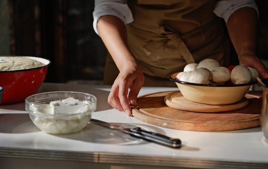 Close-up. Vintage enamel bowl with fresh mushroom champignons on wooden board and mozzarella and feta cheese on a white table against a housewife woman in a chef's apron on a rustic kitchen background
