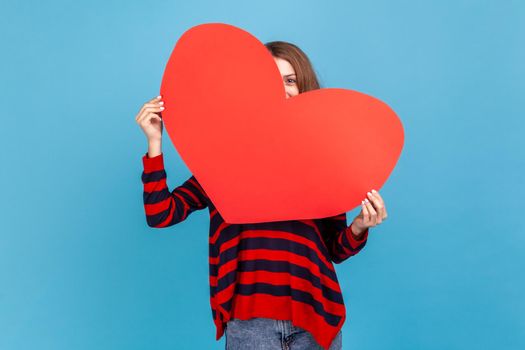 Portrait of woman wearing striped casual style sweater, peeping from big red heart, being shy to express her romantic feelings and love. Indoor studio shot isolated on blue background.