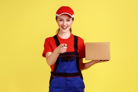 Portrait of smiling optimistic delivery woman standing with carton box, parcel for client, pointing to camera, wearing overalls and red cap. Indoor studio shot isolated on yellow background.