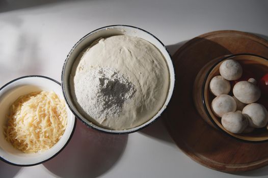 Flat lay. Enamel bowls with yeast rising dough, sprinkled with white flour, grated cheese, fresh mushroom champignons on a wooden cutting board on a table in a country cottage kitchen. Food still life