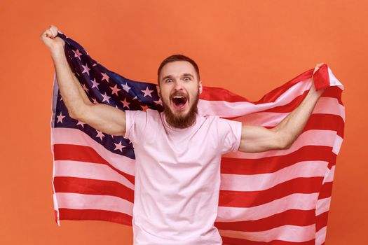 Portrait of bearded man holding USA flag and screaming happily, looking at camera, celebrating national holiday, wearing pink T-shirt. Indoor studio shot isolated on orange background.