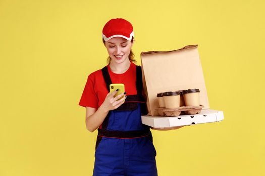 Portrait of positive delivery woman holding coffee and pizza boxes, using smart phone for checking address for delivery, wearing overalls and red cap. Indoor studio shot isolated on yellow background.