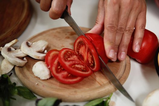Close-up. Details of the hands of a chef slicing ripe juicy tomato on a wooden chopping board with chopped mushroom champignons, while preparing healthy vegan food. Veganism and healthy eating concept
