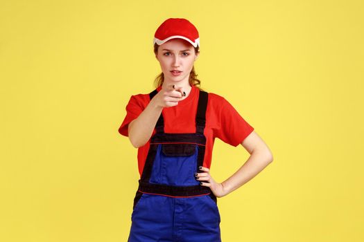Portrait of serious strict worker woman standing with hand on hip and pointing at camera with finger, wearing overalls and red cap. Indoor studio shot isolated on yellow background.