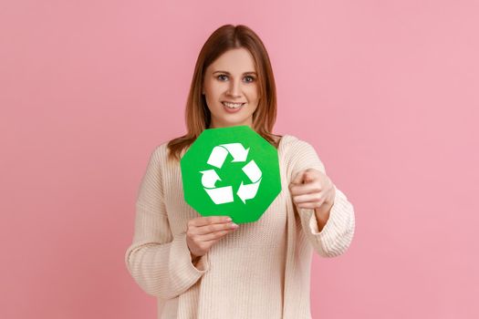 Portrait of happy satisfied young adult blond woman holding green recycling sign, saving environment, pointing at camera, wearing white sweater. Indoor studio shot isolated on pink background.