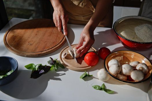 View from above of the hands of a female chef cutting mushrooms on a wooden cutting board. Freshly picked culinary herbs, organic tomatoes and raising yeast dough on an enamel bowl on a kitchen table