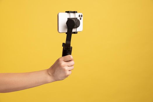 Closeup side view of woman hand holding steadicam with phone, for making video or has livestream. Indoor studio shot isolated on yellow background.