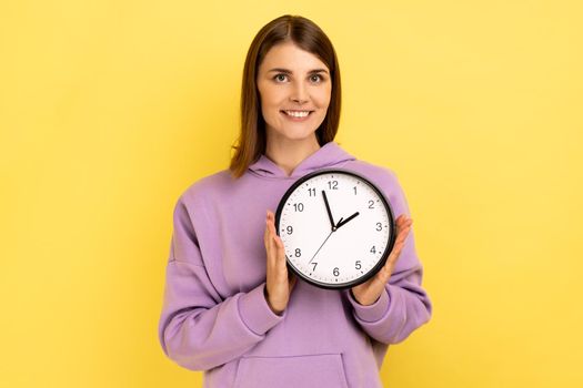 Portrait of happy positive woman holding big wall clock, looking at camera with pleasant smile, time to go, wearing purple hoodie. Indoor studio shot isolated on yellow background.