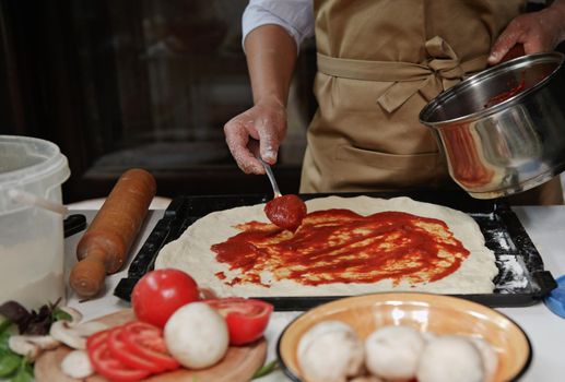 Cropped view of a housewife in beige chef's apron spreading freshly made tomato sauce on rolled out dough in pizza pan. Fresh organic ingredients on a kitchen table. Cooking. Culinary. Italian cuisine