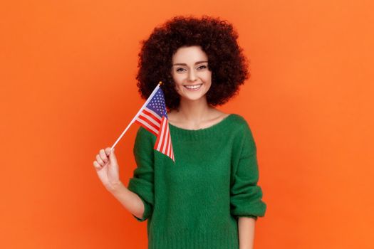 Happy positive woman with Afro hairstyle wearing green casual style sweater standing and waving american flag, celebrating national holiday. Indoor studio shot isolated on orange background.