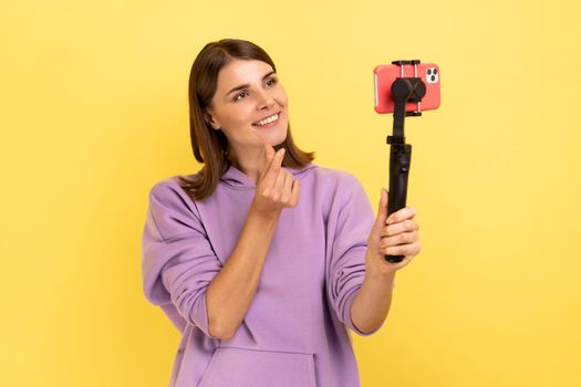 Optimistic woman using cell phone and steadicam for broadcasting livestream, showing korean finger heart sign, symbol of love, care, peace or support. Indoor studio shot isolated on yellow background.