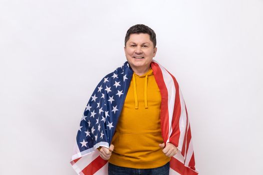 Happy relaxed satisfied handsome middle aged man standing wrapped in USA flag, celebrating national holiday, wearing urban style hoodie. Indoor studio shot isolated on white background.