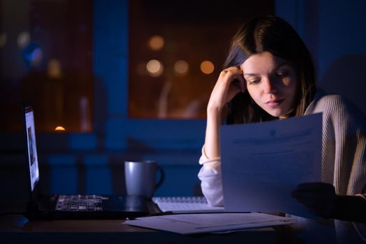 A young girl works at home with a laptop at night and talks on the mobile phone, goes through papers, documents, compiles analytics, makes calculations and notes, solves business issues, studies.