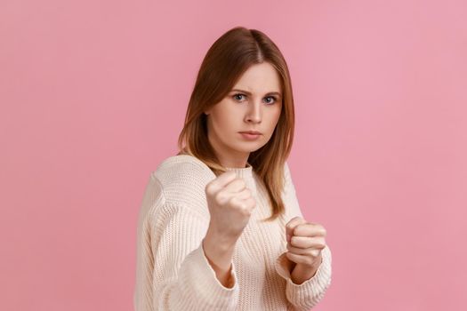 Portrait of angry confident attractive blond woman clenching fists, showing boxing gesture and ready to punch, wearing white sweater. Indoor studio shot isolated on pink background.