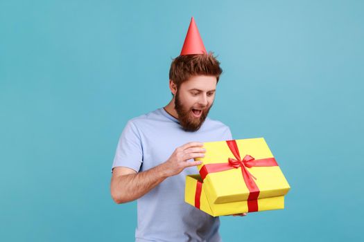 Man with party cone hat holding opened gift box and looking at present with excited look, unpacking present, enjoying awesome birthday surprise. Indoor studio shot isolated on blue background.