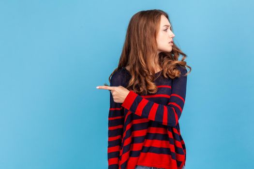 Get out. Portrait of angry resentful woman wearing striped casual style sweater, pointing way out and grimacing madly, ordering to leave, showing exit. Indoor studio shot isolated on blue background.