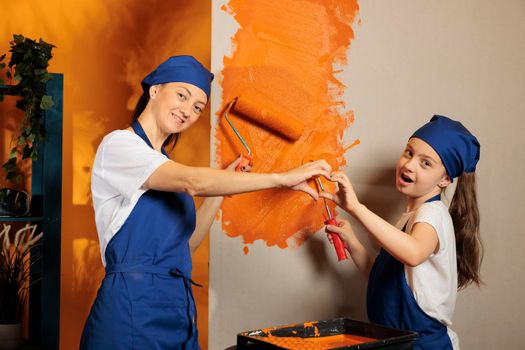 Mom and kid donig heart shape sign and using orange paint to color house walls, romantic love gesture. Painting apartment room and showing romance feelings symbol, renovating home.