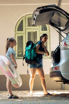 Mother with young child going on holiday with automobile, travelling to seaside with luggage and inflatable. Leaving on summer vacation, loading travel bags and trolley in vehicle trunk.