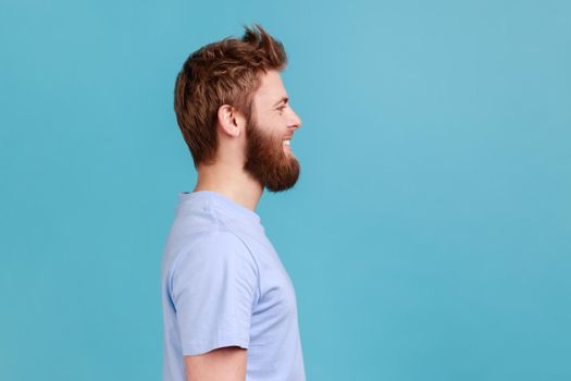 Side view of bearded man looking at camera with toothy smile and happy facial expression, being in good mood, rejoicing great results. Indoor studio shot isolated on blue background.