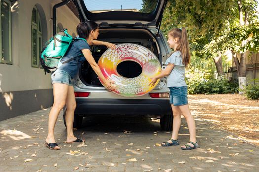 Woman and young kid travelling by car, going to seaside with inflatable and luggage. Loading suitcase, baggage and travel bags in trunk of vehicle to leave on summer holiday vacation.