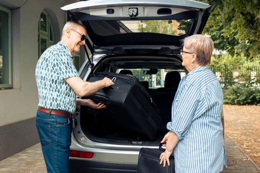 Aged couple putting bags in vehicle trunk to leave on retirement holiday trip during summer. Travelling on road trip for recreation, packing baggage and suitcase for urban cityscape vacation.