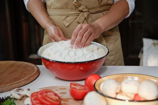 Close-up of the hands of a housewife, female pastry chef in a beige apron, kneading yeast dough for Italian vegetarian pizza, in a vintage enamel bowl in a rustic kitchen