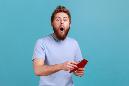 Portrait of surprised young adult handsome bearded man reading letter or greeting card, holding envelope, being astonished of shocked news. Indoor studio shot isolated on blue background.