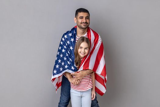 Portrait of optimistic father and daughter standing wrapped in big flag of United States of America together, celebrating national holiday. Indoor studio shot isolated on gray background.