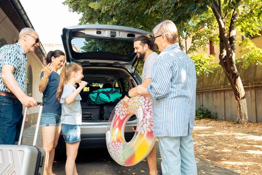 Happy people travelling at seaside with vehicle, leaving on vacation trip with parents, grandparents and small girl. Loading luggage and suitcase in automobile trunk, summer adventure.