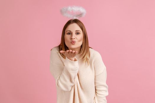 Portrait of romantic charming blond angelic woman with nimbus over head looking at camera and sending air kisses, wearing white sweater. Indoor studio shot isolated on pink background.