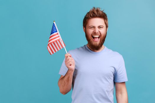 Portrait of overjoyed bearded man holding in hand flag of united states of america, celebrating independence day, having excited facial expression. Indoor studio shot isolated on blue background.