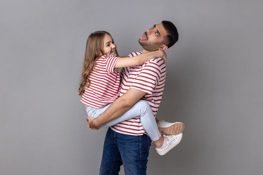 Portrait of funny father and smiling charming daughter in striped T-shirts standing together, tired dad holding kid and showing tongue out. Indoor studio shot isolated on gray background.