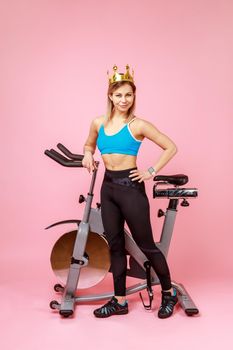 Full length of athletic woman in golden crown, queen of sports trainings, standing with hand on hips near bike simulator, wearing sports tights and top. Indoor studio shot isolated on pink background.