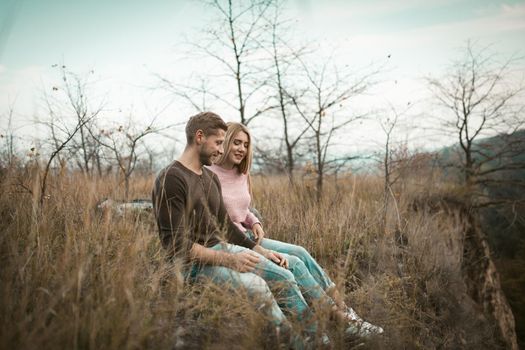 Happy Man And A Woman Are Sitting On The Edge Of A Cliff With Their Legs Dangling Down. Couple Of Tourists Sitting On The Grass On A Hillside In The Fall In The Fresh Air Outdoors