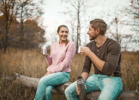 Couple In Love Drinks A Hot Drink In Nature, Focus On Caucasian Man Discussion With Woman Holding Cups Of Hot Drinks From A Thermos While Sitting On A Fallen Tree In Nature, Outdors Concept