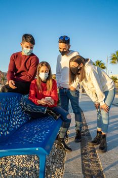 Millenial friends using smart phones wearing protective face mask on Covid wave. Worried guys and girls watching news on mobile smartphone. Travellers group stay in touch during pandemic in vacations