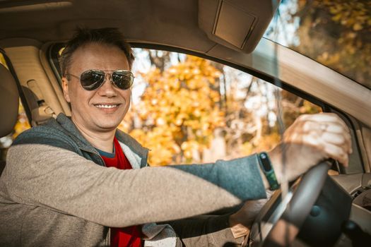 Driver stopped car in autumn forest. Handsome man in sunglasses smiles at the camera sitting in the car against the background of trees with yellow foliage outside the window.