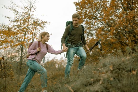 Loving couple of tourists climb the slope holding hands. Cheerful Young Couple, man and a woman with backpacks travel together in autumn nature. Hiking concept. Healthy lifestyle concept.