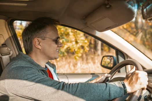 Male Driver Driving A Car, Side View Of Smiling Caucasian Man In Sunglasses Holds Steering Wheel Of Car While Sitting In Light Interior In Beige Color And Looking At Road, Road Trip In Nature