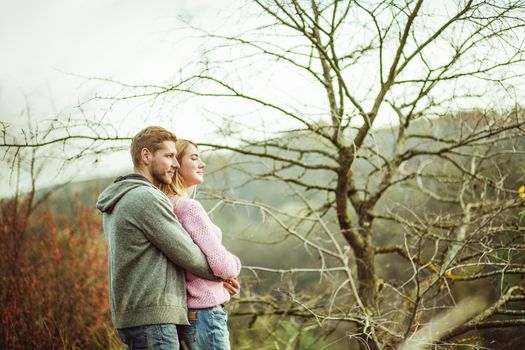 Hugging couple stands on top of hill admiring the sunset. Against the background of a tree without leaves. Autumn mood concert.