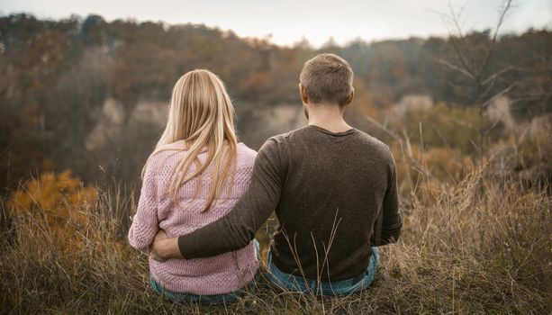 Couple Sits Embracing On Cliff Edge, Rear View Of Couple In Love Admiring Sunset Outdoors, With Autumn Forest And Cloudy Sky On Blurred Background