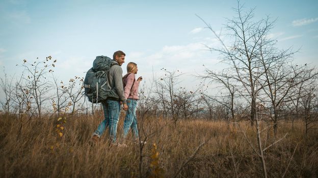 Couple of travelers walking through an autumn field holding hands. Tourists at full height against a background of blue sky and autumn natural landscape. Shot from below. Hiking concept.