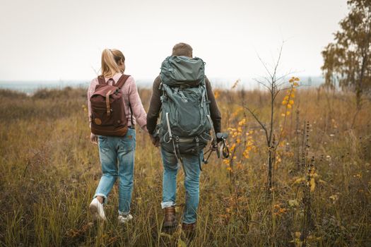 Hiking in autumn nature, couple of backpackers makes their way across the field, rear view of man and woman with backpacks and hiking poles walking in nature outdoors. Hiking concept.