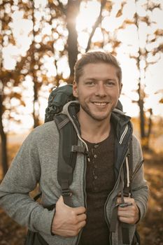 Male Backpacker Hiking In Autumn Forest, Portrait Of Young Caucasian Man In Gray Jacket Looking At Camera While Walking Towards New Adventures In Nature On Sunny Day Outdoors