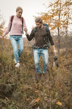 Two tourists are walking on the hill holding hands, a man is supporting a beautiful blonde with a backpack walking together in the wild nature in autumn. Hiking Concept.